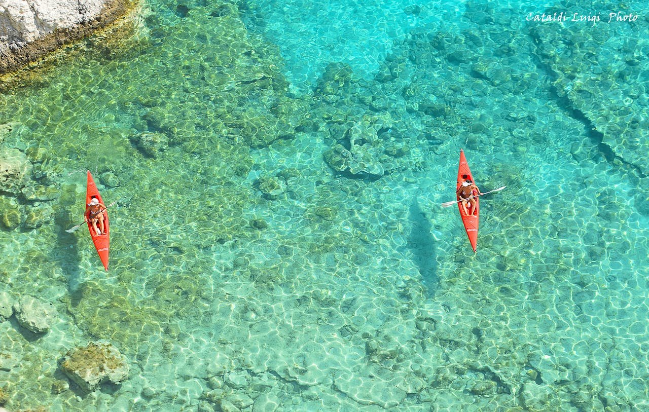 Aerial view of two kayakers paddling on crystal clear turquoise water near a rocky shore.
