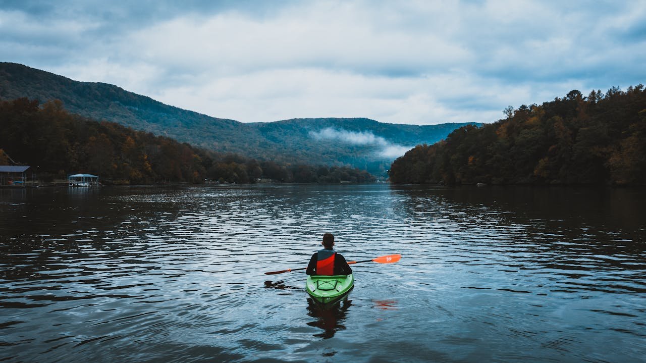 A lone kayaker paddles on a calm, misty lake surrounded by scenic mountains and autumn trees.